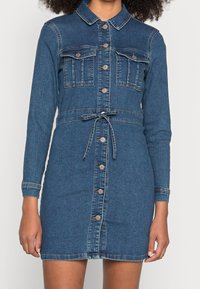 Rikka deniimi, jossa on pitkät hihat, kaulus, kaksi rinta-taskua, säädettävä vyötärönauha ja napitus edessä. Tumma sininen väri ja sileä pinta.