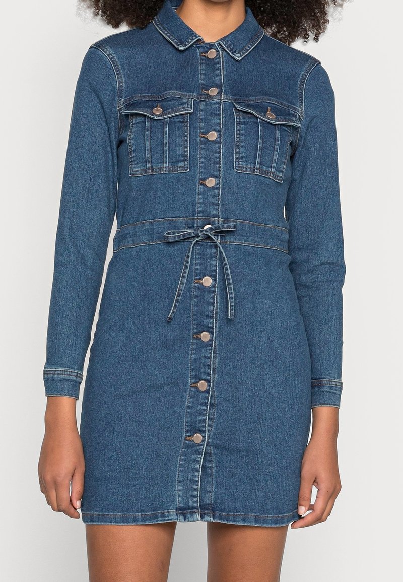 Rikka deniimi, jossa on pitkät hihat, kaulus, kaksi rinta-taskua, säädettävä vyötärönauha ja napitus edessä. Tumma sininen väri ja sileä pinta.