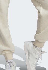 Person som bär beige sweatpants och vita sneakers, med en fot något upphöjd som visar skosålen.