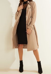 Manteau beige avec des boutons argentés, manches longues et taille ceinturée sur une robe noire. Le modèle porte des bottines noires. Texture lisse.