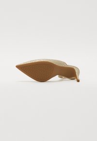 Chaussure à talons hauts dorée scintillante avec un bout pointu et une semelle brune texturée. Présente un talon fin et une finition métallique sur toute la surface.