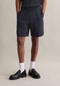 Marineblauwe shorts met een elastische tailleband, voorzien van twee zijzakken; gedragen met zwarte instapschoenen en witte sokken. Gestructureerde stof.