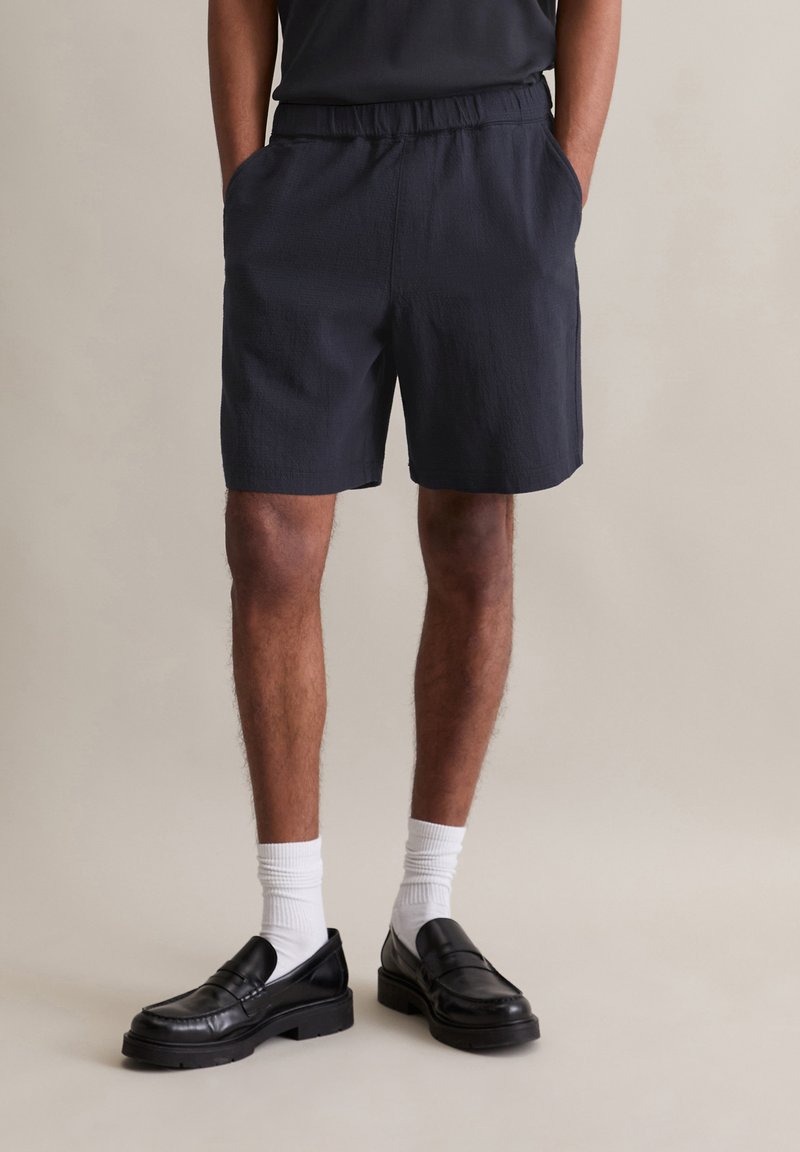 Marineblauwe shorts met een elastische tailleband, voorzien van twee zijzakken; gedragen met zwarte instapschoenen en witte sokken. Gestructureerde stof.