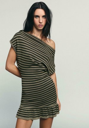 Žena s dlhými tmavými vlasmi, oblečená v krátkych olivovo-čiernych pruhovaných šatách s odhalenými ramenami a volánikovou spodnou časťou, stojaca pred jednoduchým pozadím.