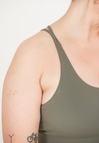 Oliivinvihreä urheiluliiivi ohjeilla, jossa on sileä pinta jaulotettu muotoilu, joka paljastaa osittain tatuoinnin yläkäsivarressa.