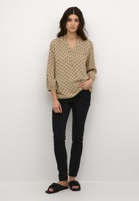 Een tan blouse met zwarte stippen, een v-hals, en ballonmouwen, gecombineerd met donkere skinny jeans en zwarte instapsandalen.