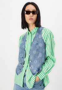 Denim vest with floral detailing, worn over a green and white striped button-up shirt. Features a fitted design and front buttons.