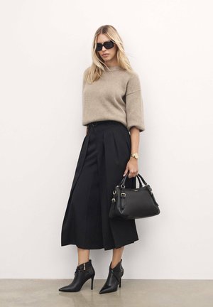 Suéter de punto beige, culottes plisados negros, bolso negro, botines cortos negros de punta. La modelo posa contra un fondo neutro.