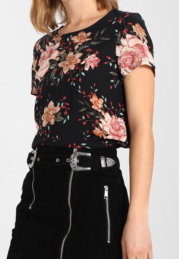 Femme portant un haut noir à manches courtes à motif floral, associé à une jupe noire avec des détails de fermeture éclair argentés et une boucle de ceinture décorative.