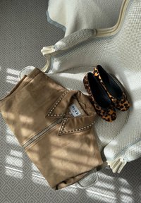 Chaqueta de ante sintético marrón con tachuelas plateadas en el cuello, combinada con bailarinas negras con un estampado de leopardo. Colocada sobre una silla texturizada.