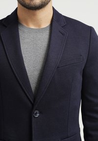 Navy jacket with a structured design, featuring a single button closure and a chest pocket, layered over a light grey sweater.