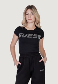 Musthave mustaviljad musta värvi lühike topp, millel on hõbedased rhinestone'ide kirjad "GUESS", koos mustade sportlike pükstega, millel on samas logoga tasku.