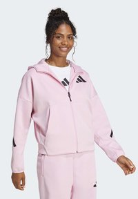 Felpa con zip di colore rosa chiaro, realizzata in tessuto morbido, che presenta una vestibilità comoda, dettagli in logo nero e cappuccio. Abbinata a pantaloni coordinati.