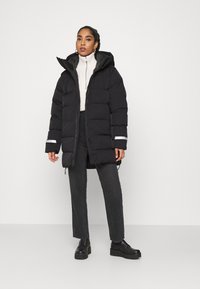 Abrigo puffer negro con capucha, con cierre de cremallera y detalles reflectantes, usado sobre un suéter ligero y pantalones oscuros, combinado con botas negras.