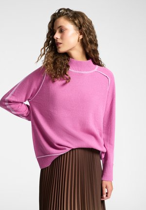 Różowy dzianinowy sweter z wysokim kołnierzem, prążkowanymi mankietami i kontrastowymi szwami. Noszony na plisowanej brązowej spódnicy, co dodaje warstwowej tekstury.