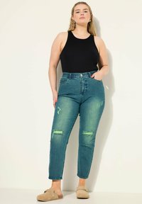 Tmavě modré džíny s vysokým pasem a roztrhanými nášivkami, spárované s černým topem bez rukávů. Džíny mají hladkou texturu a klasický design s pěti kapsami.