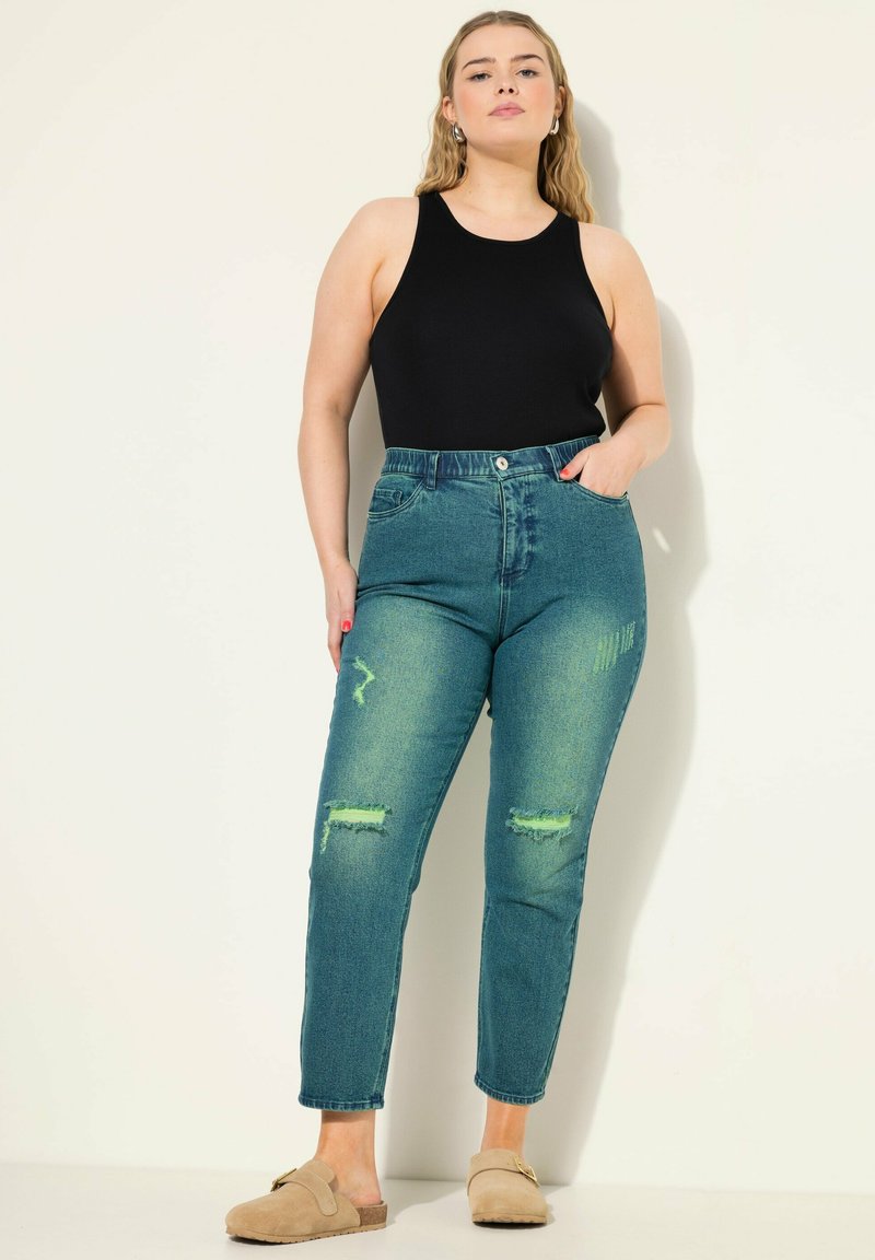 Tmavě modré džíny s vysokým pasem a roztrhanými nášivkami, spárované s černým topem bez rukávů. Džíny mají hladkou texturu a klasický design s pěti kapsami.