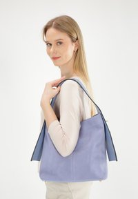 Lilla ruskind skuldertaske med en glat overflade, der har en struktureret design og kontrasterende marineblå håndtag. Messingdetaljer i bunden.