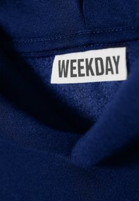 Temno modra tkanina z mehkimi teksturami in vidno šivanim. Ima belo oznako z besedilom "WEEKDAY" v debelem črnem tisku.