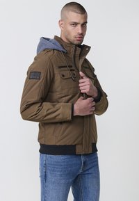 Braune isolierte Jacke mit grauem Kapuzenpullover, ausgestattet mit einem Reißverschluss vorne, mehreren Taschen und gerippten Bündchen für eine enge Passform.