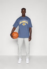 Prevelika siva majica s grafikonom "Los Angeles Lakers", v kombinaciji s sivimi trenirkami in belimi športnimi copati, drži košarkarsko žogo.