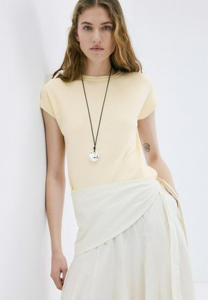 Jeune femme aux cheveux ondulés portant un haut à manches courtes jaune pâle et une jupe portefeuille blanche, debout contre un mur blanc.