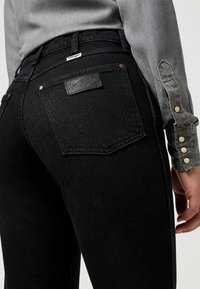Vaqueros de mezclilla negra con cintura alta, que cuentan con bolsillos traseros, una etiqueta de marca de cuero y detalles de costura sutiles. Textura suave.