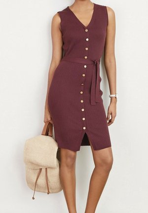 Robe bordeaux sans manches à côtes avec des boutons dorés sur le devant et une ceinture, portée par une personne tenant un sac à dos beige texturé avec des bretelles en cuir.