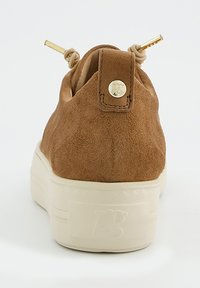 Baskets en daim marron avec une pointe arrondie, une semelle crème, des accents dorés sur les lacets et un patch logo au talon. Texture lisse dans l'ensemble.