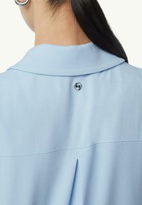 Jasnoniebieska koszula o teksturowanej tkaninie, z kołnierzykiem i małym detalem logo z tyłu, blisko dekoltu.