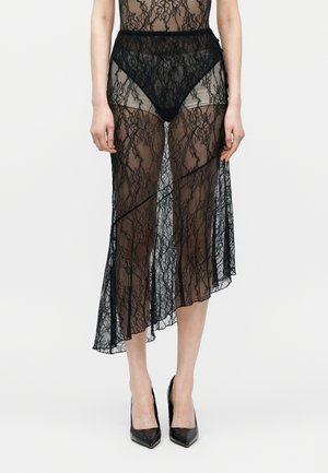 Γυναίκα που φοράει μαύρη διαφανή φούστα από δαντέλα με ασύμμετρο τελείωμα πάνω από μαύρα σλιπ με ψηλή μέση και μαύρες μυτερές γόβες.