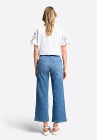 Donna con lunghi capelli biondi che indossa una blusa bianca a maniche corte con fiocchi, jeans blu a vita alta e gamba larga, e sandali bianchi, in piedi di spalle.