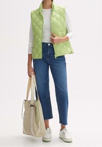 Jasnozielona pikowana kamizelka z wysokim kołnierzem, zestawiona z niebieskimi spodniami o prostym kroju i białymi trampkami. Jasnobeżowa torba na sznurek z zielonymi akcentami.