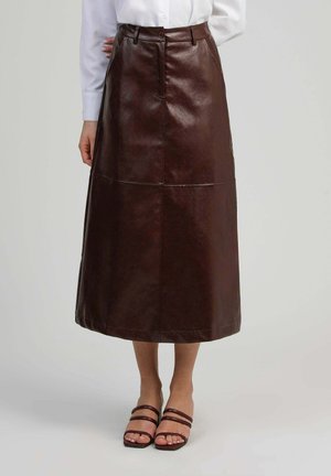 Femme portant une jupe midi en cuir marron avec des détails de couture à l'avant, associée à une chemise blanche boutonnée et des sandales marron à brides.