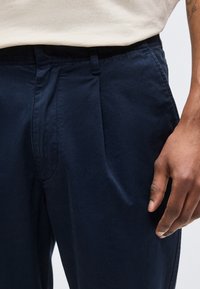 Άνδρας που φοράει μπλε σκούρο παντελόνι και λευκό πουκάμισο, με το δεξί χέρι χαλαρό στο πλευρό του και ένα τατουάζ ορατό στο αντιβράχιο.