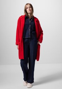 Röd oversize fleecejacka över en mörk denimskjorta och vida jeans. Lätta vita sneakers fulländar outfiten med mjuka material.