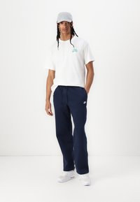 Vit bomulls-T-shirt med grönt Nike-logotyp, marinblå fleecemjukisbyxor, grå keps och vita sportiga sneakers. Avslappnad outfit med ledig passform.