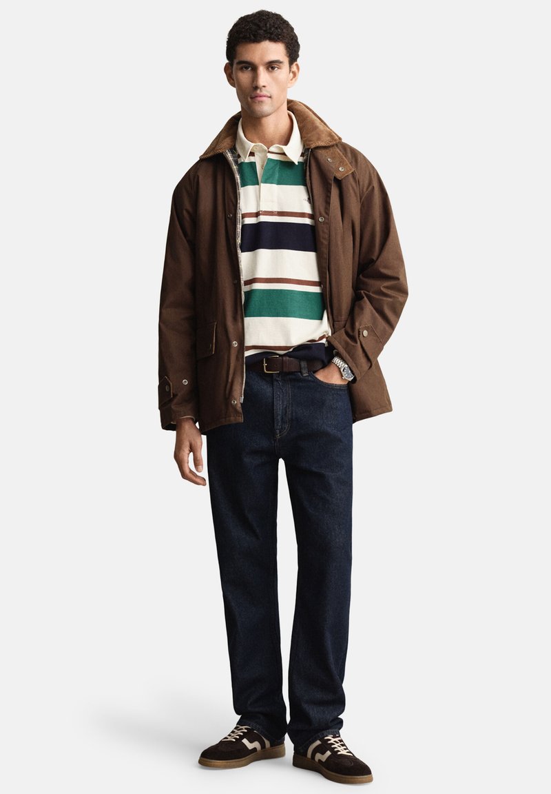 Brown jacket with a tan collar over a multi-striped polo shirt in green, white, and navy. Dark jeans and brown sneakers complete the outfit.