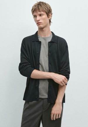 Giovane uomo con capelli biondi corti che indossa un cardigan nero con bottoni sopra una maglietta grigia, mentre si sistema la manica sinistra su uno sfondo chiaro e uniforme.