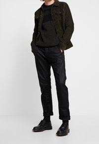 Zielona kordurojowa kurtka na czarnej swetrze, w zestawieniu z czarnymi zwężanymi spodniami i czarnymi sznurowanymi butami. Zwięzłe, nowoczesne szczegóły stroju.