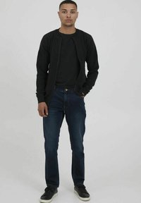 Svart zip-up jacka över en svart t-shirt, i kombination med mörkblå jeans. Modellen bär svarta sneakers och står mot en enfärgad bakgrund.