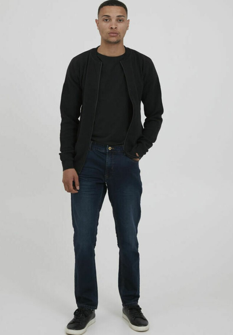 Svart zip-up jacka över en svart t-shirt, i kombination med mörkblå jeans. Modellen bär svarta sneakers och står mot en enfärgad bakgrund.