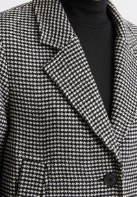 Blazer de patrón de jaquard en blanco y negro con solapa de pico, cierre de botón y detalle de bolsillo frontal, fabricado en tejido texturizado.