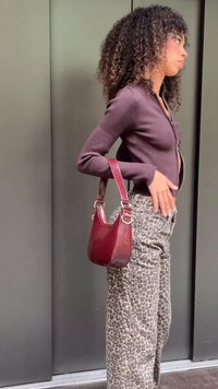 Geantă lucioasă burgund, cu formă rotunjită și curea reglabilă, asortată cu un top și pantaloni gri, strâmți, cu imprimeu leopard și model reiat.