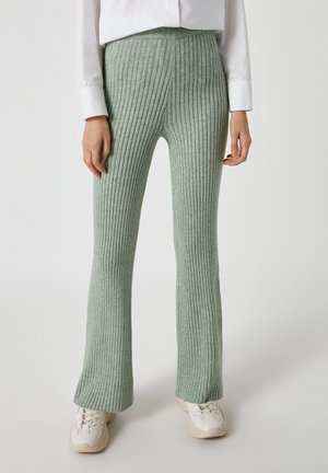 Modelo que lleva pantalones acampanados de punto acanalado verde claro con zapatillas blancas y una camisa blanca de manga larga, de pie sobre un fondo liso.