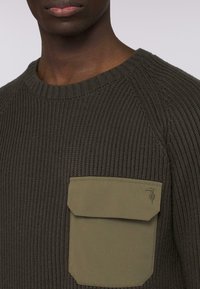 Tmavě zelený pletený svetr s ribbovanou strukturou, kulatým výstřihem a velkou olivově zelenou náprsní kapsou na levé straně. Detail loga na kapse.