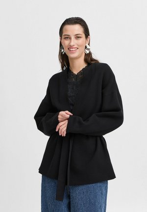 Cardigan negru tricotat cu mâneci largi, design înfășurat și talie legată. Stratificat peste un top din dantelă neagră, asortat cu pantaloni cu dungi albastre.