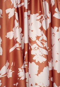 Samtstoff in Terrakotta mit großen weißen Blumenmustern; glatte Textur mit drapierten Falten, die den lebhaften Farbkontrast betonen.