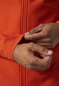 Chaqueta naranja con cremallera y tejido texturizado, con un primer plano de manos ajustando el puño. Detalles de costura y cremallera visibles.