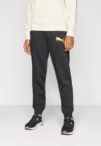 Sorte joggebukser laget av mykt stoff, med en gull Puma-logo på venstre lår. Kombinert med svarte joggesko med rosa detaljer.